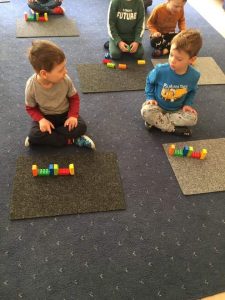 Na dywanie siedzi kilkoro dzieci, a przed nimi leży zestaw 6 klocków LEGO ustawiony w identycznej konfiguracji jaką zaprezentował nauczyciel.