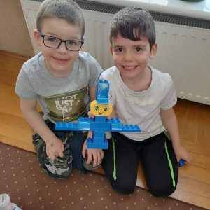 Dwóch chłopców siedzi na podłodze i prezentuje model niebieskiego ludzika z klocków LEGO.