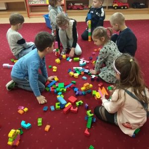 Na dywanie siedzą dzieci wokół rozrzuconych klocków LEGO i budują z nich różne konstrukcje.