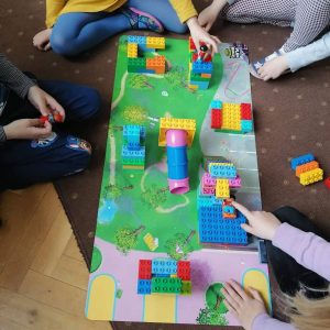 Na macie przedstawiającej krajobraz zieleni dzieci budują z klocków LEGO plac zabaw.