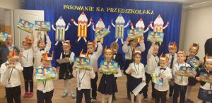 Dzieci ubrane w odświętne stroje, unoszą w górę kolorowe dyplomy, które dostały z okazji Pasowania na Przedszkolaka.