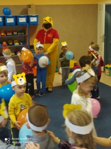 Dzieci ubrane w kolorowe opaski z wizerunkiem misia, trzymają w rękach kolorowe balony. Wśród nich jest osoba przebrana z Misia Puchatka.